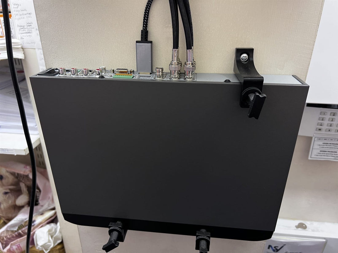 Grey CCTV DVR held on a wall by three separate adjustable brackets, the top bracket screwed to the wall beside the connectors